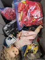 Lot contents showing a vintage doll, toy vehicles and bags of red building blocks inside a storage bin