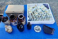 Top-down view of vintage ceramic and glass jugs, ashtrays, and boxed unused glasses shown on blue surface