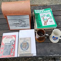 Top-down view of large folder with helicopter photo inside cover, old car flyers/magazine cutouts, two mugs and a stack of vintage Stamp Magazines