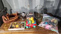 Various items arranged on wooden table showing wooden bowls, hand blown pitcher, toy car with duck figurines, CDs, cassette tape box set, and a small metallic bowl.