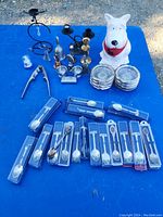 Photo showing white ceramic dog cookie jar, glass coasters in stacks, multiple sets of boxed collectible spoons, black metal candle holders, small glass bell, nutcracker, and other small items arranged on blue surface.