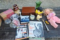 Wide view of items spread on a wooden surface showing the doll, wooden boxes, magazines, glassware, yellow bust, ceramic jug, wrench, sharpener, and small toy car.