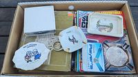 Photo of box with car magazines, three ashtrays including one with a cartoon couple, a tin of stamps, and a Zippo branded belt