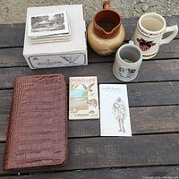 Photo showing vintage travel kit, ceramic pitcher, two mugs, and stacks of vintage postcards on a wooden surface
