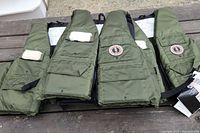 Three olive green survival sports vests arranged side-by-side on a wooden surface, each with four pockets and visible logo patches.