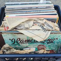 Top view of plastic crate filled with numerous vinyl records and some old newspaper and paper liners visible.