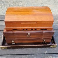 Three wooden items arranged on a dark wood surface: a wood tray base, a wood box with two drawers stacked on the tray, and a curved wood jewelry box stacked on top of the box.
