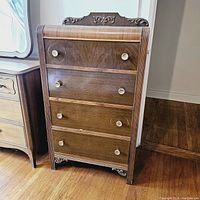 Front view of antique solid wood tall dresser showing four drawers and decorative carved top and feet.