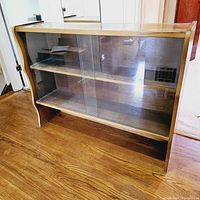 Front and side view of a vintage Mid Century Modern solid wood display case with glass sliding doors and two internal shelves, placed on wooden floor.
