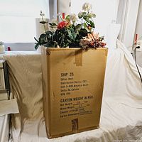 Tall cardboard box filled with assorted vintage artificial flowers and floral arrangements.