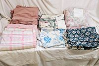 Full overview of the lot showing folded bed sheets sorted in stacks by color and pattern including solid dusty pink, pastel stripes, large floral prints, and navy blue geometric floral patterns.