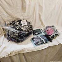 Wide shot of a large transparent plastic sack filled with various folded scarves, alongside three smaller sealed plastic bags with additional scarves inside, displayed on a white fabric backdrop.
