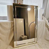 Full view of the large vintage brass mirror leaning against a white surface, showing the rectangular shape and reflective glass with brass frame edges.