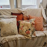 Wide shot showing eight pillows: two vintage white hand sewn pillows sealed in plastic, four rust-colored pillows, two floral patterned pillows with soft multicolor design.