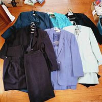 Multiple vintage 2 piece suits laid out showing variety of colors and fabrics including navy, black, teal, light lavender, and mint green.