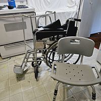 Photo showing the shower chair support in front, with black and silver wheelchair and metal walker behind it.