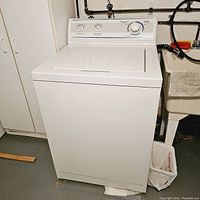 Front angle view of a white Magic Chef washing machine placed on a basement floor, white rectangular lid closed, showing minor wear and rust at bottom front.