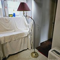 Full view of the tall floor lamp showing the brass base, curved dark stem, and burgundy pleated shade.