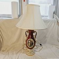 Full view of the lamp with white fabric shade and ceramic base with painted artwork and cord visible.