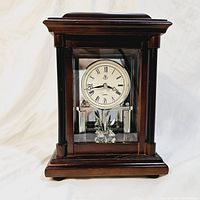 Front view of dark mahogany Woodpecker mantel clock showing glass front panel, Roman numeral clock face, and crystal pendulum inside.