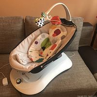 Side view of 4moms Rockaroo infant swing showing beige seat with colorful padded insert and control dial on base.