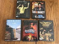 Five music-related DVD cases arranged on a wooden surface showing Shania Twain, Sid & Nancy, Bon Jovi, Jimi Hendrix, and Aerosmith covers.