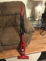 Red Dirt Devil Swift Stick vacuum cleaner placed on a brown corduroy armchair, showing the full length of the stick with handle and vacuum base.