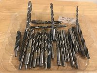 Top view of multiple large size metal drill bits arranged on a flat surface, showing various drill bit sizes and fluted metal shafts.