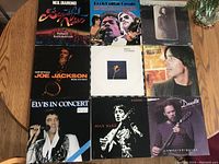 Top-down photo of nine LP vinyl records from various artists arranged on a wooden surface. Shows minor surface wear such as light scratches and scuffs on album covers.