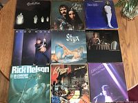 Photo showing nine vinyl LP albums laid flat on wooden table, arranged in three rows of three. Album covers visible with various musical artists and titles.