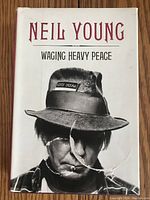 Front cover of the hardcover book showing the title 'NEIL YOUNG WAGING HEAVY PEACE' with an image of Neil Young wearing a hat with a patch that reads 'Some Dream'. The dust jacket has a significant tear near the lower center.