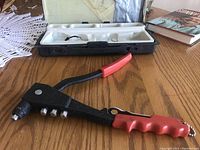 Side view of the handheld riveter tool with black metal body and red handles laying next to its carrying case with white molded insert.