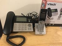View of the Vtech corded phone base with digital display and keypad, alongside a cordless handset on charging base, with original packaging visible in background.