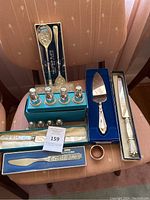Photo showing multiple silver serving items on a chair including boxed salt and pepper shakers, cake server, butter knife, and silver spoons.