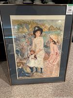 Framed art print of children on the seashore, displayed upright on carpeted floor. The image shows four children dressed in vintage attire near a wooden fence with water and green trees behind.