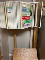 Photo showing white multi-panel metal display frame open with various colorful printed humorous and satirical signs attached, mounted on a wooden stand base. Some example signs include 'I'm Always in the Ozone I Come Everywhere,' 'High on Stress & Low on Cash,' and 'SWF Seeks $$$.'