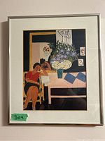 Frontal photo of framed artwork depicting a woman reading beside a table with flowers, with visible details of composition and colors.
