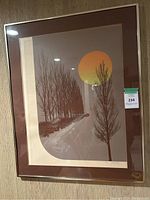 Framed MCM style art print with brown matting and gold metallic frame, showing a stylized winter landscape with tall bare trees and a large orange circular sun or moon.