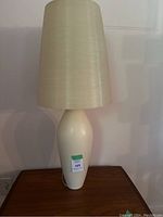 Front view of a cream colored ceramic table lamp with tall conical plastic shade on wooden surface
