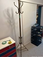 Photo of a tall metal coat hanger with upper and lower hooks, tripod base, placed against a white wall.