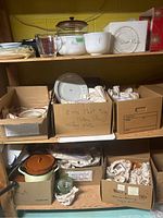Shelves showing various kitchen items including glassware, plates in cardboard boxes, labeled meat platters, bowls, and mixing dishes.