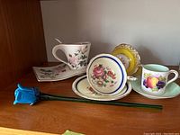 Wooden shelf displaying four fine china teacups and saucers with different floral and fruit patterns, alongside a blue glass rose decoration.