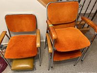 Three retro cantilever chairs with orange plush seats and padded wooden armrests, arranged together indoors.