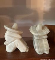 Pair of carved stone bookends shaped as seated human figures wearing sombreros, captured on a wooden shelf with warm lighting highlighting stone texture and coloring.