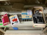 Shelf with Argus movie camera in box, blue and white box for camera light labeled Acme-Lite, and several small cleaning items.