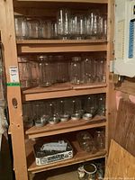 Wooden shelving unit holding multiple clear glass canning jars on top three shelves and some jars and lids on bottom shelf, showing various sizes and shapes of jars.