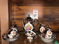 Full view of entire coffee set including teapot, sugar bowl, creamer, cups and saucers with silver-tone reflective finish and romantic scenes