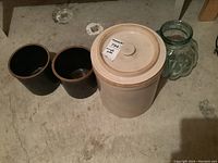 Photo of four items on concrete floor: large beige stoneware crock with lid, two smaller brown glazed redware crocks, and clear art glass jar.