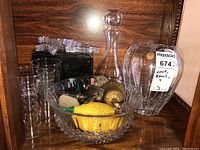 Items displayed on wooden shelf including crystal bowl with scalloped edges holding Murano glass fruit, clear glass decanter with stopper, several votive candle holders, fish figurine, and plastic frame displays.