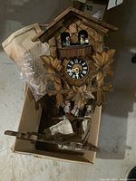 Front view of wooden cuckoo clock with carved leaves and figurines on balcony, showing clock face with Roman numerals.